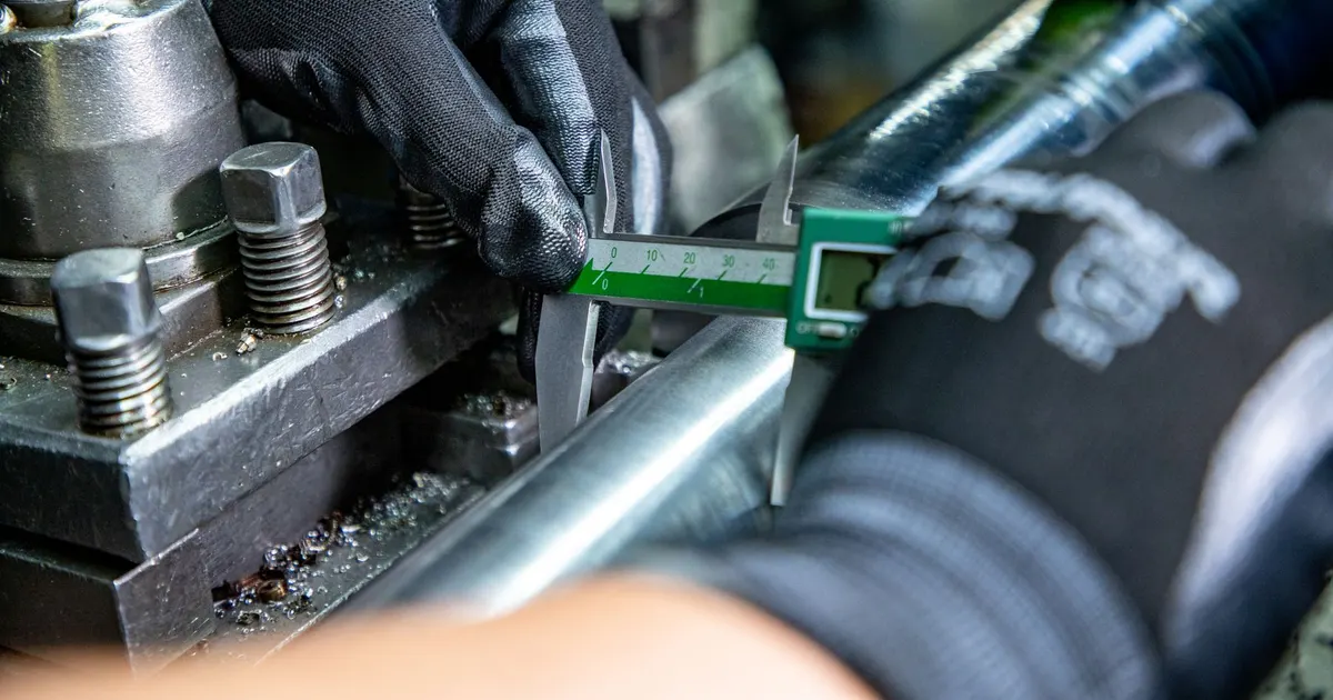 Gloved hand using a digital caliper to measure a tube held in a lathe, an example of dimensional inspection on a machined part