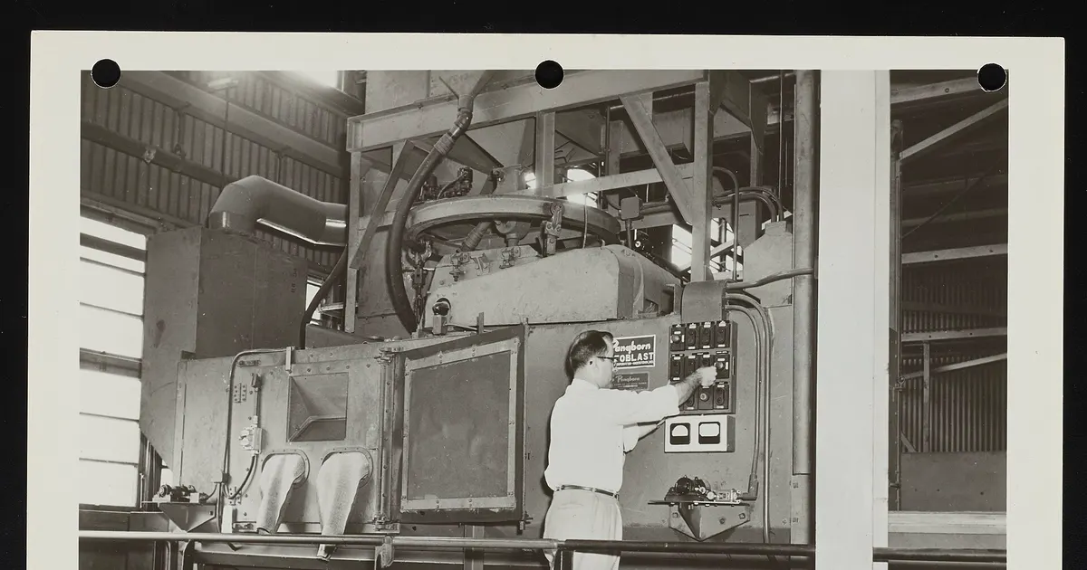 Archival photograph of an operator at industrial shot peening equipment used for fatigue-critical metal parts
